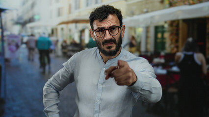 Hispanic man with beard pointing outdoors in lively street scene wearing glasses and gray shirt...