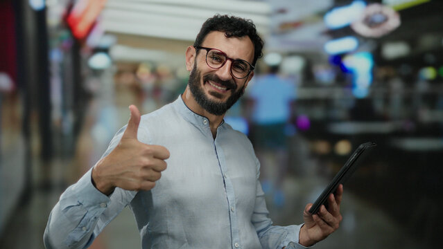 Hispanic man with beard smiling in a shopping mall while holding a tablet, engaging with technology in a vibrant retail environment. - Powered by Adobe