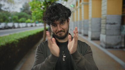 Young hispanic man with beard clapping outdoors on urban street with trees and cars in background,...