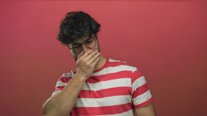 Young man with beard and glasses covering nose due to bad odor against vibrant pink background...