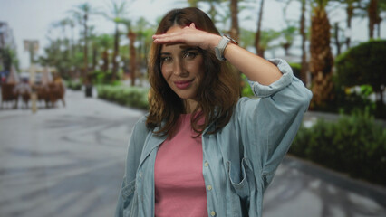 Young woman standing in a sunny park shielding her eyes with her hand, surrounded by palm trees and...