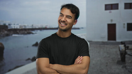 Obraz premium Young hispanic man smiling confidently on seaside promenade with arms crossed, serene ocean backdrop enhancing outdoor setting.