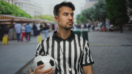 Young hispanic man in referee uniform holding soccer ball stands confidently in bustling outdoor...