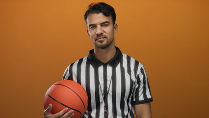 Young hispanic man wearing referee uniform holding basketball against orange background embodying...