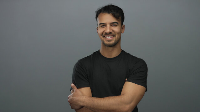 Young hispanic man smiling confidently against isolated grey background in casual black shirt showcasing friendly demeanor and positive attitude