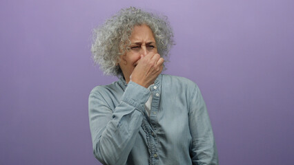 Elderly woman with gray hair appears disgusted by a smell while standing against a purple wall...