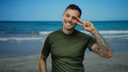 Smiling man with tattoos making peace sign at seaside with ocean in background, enjoying beach day under sunny sky, exuding happiness and relaxation.