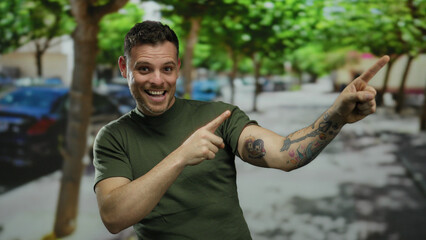 Smiling man points in street surrounded by cars and trees displaying tattoos and wearing green...