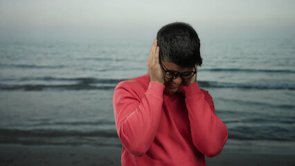 Man on beach with eyes closed covering ears wearing glasses and red shirt by the sea under overcast...