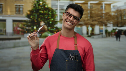 Hispanic man wearing glasses and apron holding scissors outdoors on a city street with a festive...