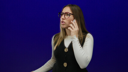 Fototapeta premium Woman talking on phone against vivid blue background, demonstrating business attire and expression, suggesting communication and technology themes.
