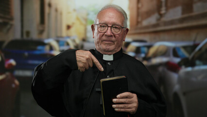 Elderly priest holding a book gestures while standing on a busy street lined with cars, implying a...