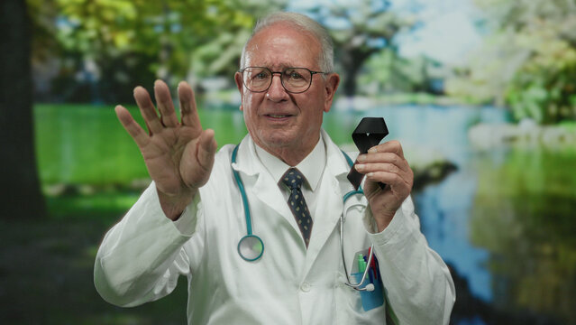 Senior male doctor in white coat standing outdoors in a park holding a black ribbon while gesturing with his hand, surrounded by lush green scenery.