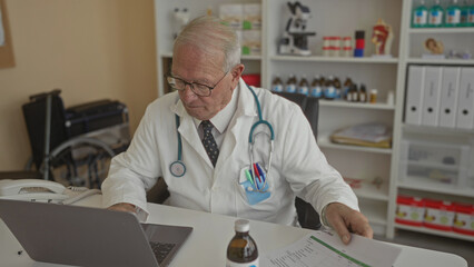 Senior male doctor in white uniform reviews paperwork in hospital room with medical equipment and laptop on desk, focused and professional.