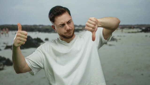 Young man with beard gives thumbs up and thumbs down at seaside beach with rocks and overcast sky, expressing mixed emotions outdoors