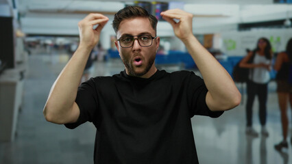Young man with beard wearing glasses gesturing in an indoor airport terminal, surrounded by...