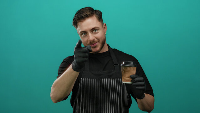Young man with beard wearing apron and gloves holds coffee cup while pointing at camera against green background, depicting a friendly waiter inviting interaction in a cafe setting.