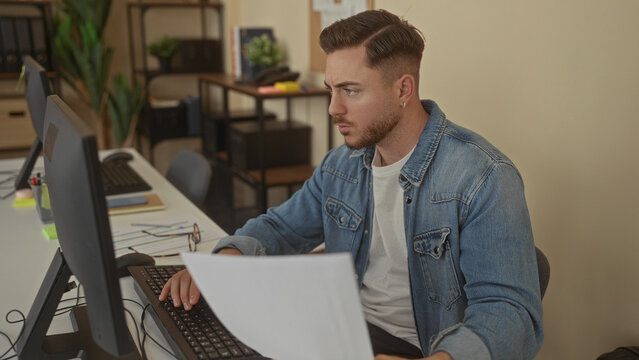 Young man with beard in denim jacket focused on work at office desk, reviewing documents by computer in modern workplace interior with shelves and plants. - Powered by Adobe