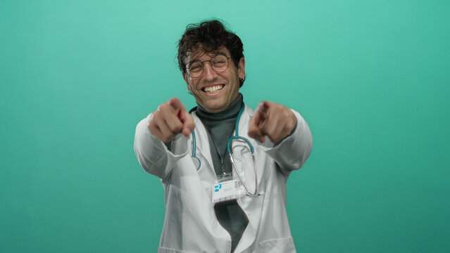 Hispanic doctor with glasses and stethoscope pointing at camera against vibrant green background, smiling confidently.