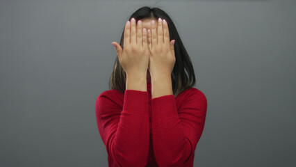 Smiling young chinese woman in red sweater reveals her face from behind hands against a plain grey...