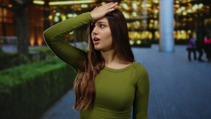 Woman in green shirt expressing confusion on city street at night with blurred lights in...