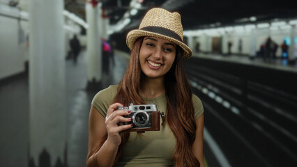 Young hispanic woman holding camera at indoor train station in hat and casual attire with railway in background, smiling, tourist ambiance captured vividly.