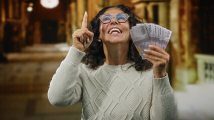 Middle-aged woman holding rupiah banknotes indoors at a church, expressing excitement and pointing...