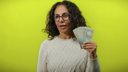Middle-aged woman holding uae dirhams with a surprised expression against a vibrant yellow...
