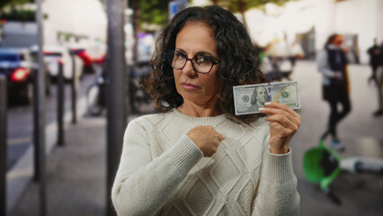 Middle-aged woman in city street holds us dollars, pointing to her heart, symbolizing financial...