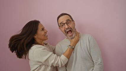 Middle-aged hispanic couple playfully interacting against a pink background, with the woman gently holding the man's neck, exuding love and humor.