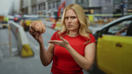 Woman holding brain in construction site, looking confused amidst busy city backdrop with traffic.
