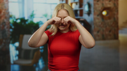 Woman indoors mimicking crying gesture in casual red outfit at home, with bookshelves in soft focus...
