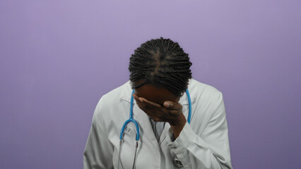 Female doctor with braided hair stands in front of a purple wall covering her face with her hand, wearing a white coat and a stethoscope suggesting stress or fatigue.