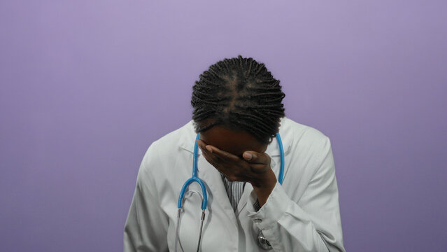 Female doctor with braided hair stands in front of a purple wall covering her face with her hand, wearing a white coat and a stethoscope suggesting stress or fatigue.
