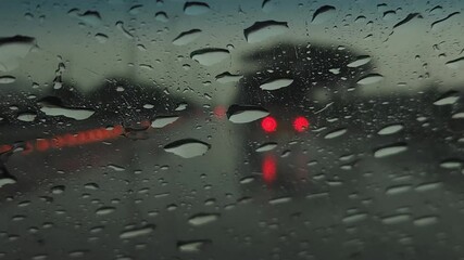 A close-up view through the rain-soaked west glass window of a motor vehicle reveals blurred lights, passing cars, and traffic reflections, as windshield wipers sweep on glass under falling rain drops - Powered by Adobe