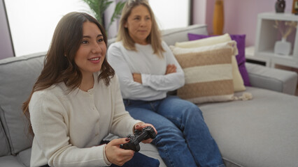 Woman playing video game while sitting on a sofa in a cozy living room with her mother watching...