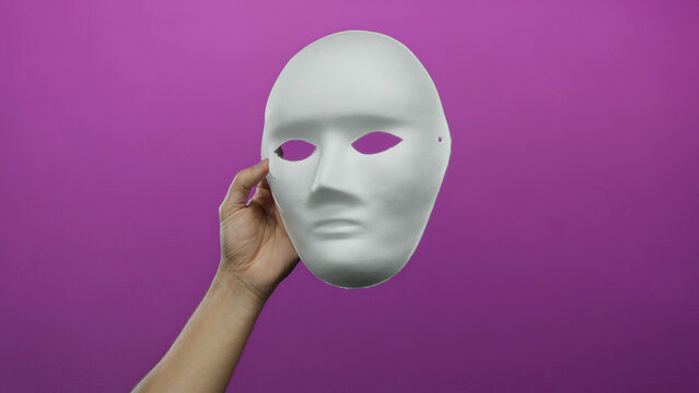 Man holding a blank white mask against a vibrant pink background, showcasing elements of anonymity and creativity isolated in a studio shot.