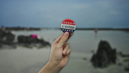 Hand of caucasian man holding an i voted badge at a seaside beach, with a focus on election...