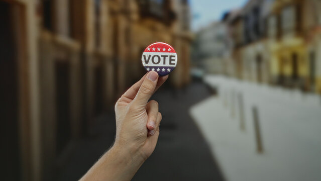 Caucasian man holding a vote badge in an outdoor city street, highlighting themes of democracy and civic engagement in an urban environment.