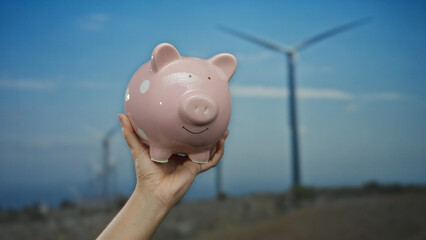 Caucasian man holds a pink piggy bank against a backdrop of windmills in an outdoor field,...