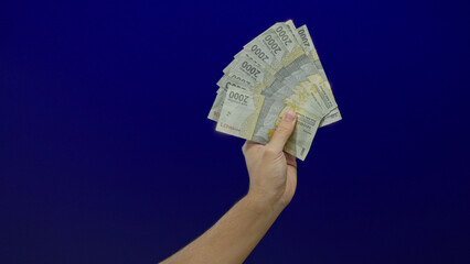 Male hand holding indonesian rupiah banknotes against an isolated blue wall background, emphasizing...