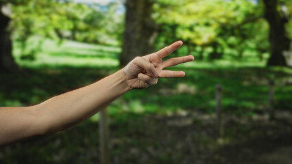 Man's hand gesturing three fingers outdoors in a lush green park setting, emphasizing nature and relaxation.