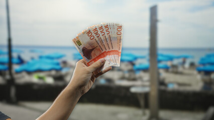 Man holding norwegian kroner banknotes at a seaside beach with blue umbrellas and water in the background, capturing a sense of travel and finance.