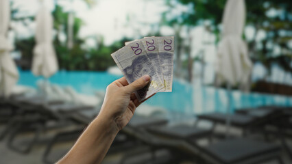 Hand holding swedish krona banknotes by a pool at a resort, illustrating financial freedom and leisure in a tropical outdoor setting, highlighting vacation expenses.