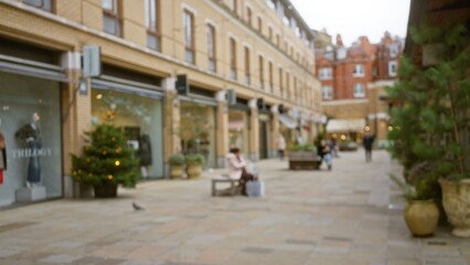 Obraz premium Woman sitting on a bench in a blurred london street scene during winter, capturing the essence of urban life in the united kingdom with defocused architectural background.