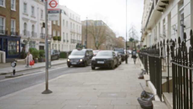 Blurred view of a london street in autumn with a woman carrying a red bag, cars parked along the sidewalk, and soft focus buildings in the background.