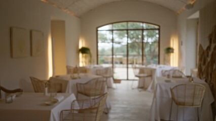 Blurred view of elegant restaurant interior with defocused tables and chairs, featuring soft lighting and bokeh effect through large windows.