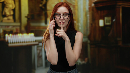 Young redhead woman gestures silence in a church setting, conveying discretion and calmness with...