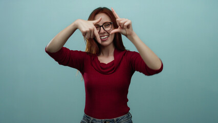Smiling woman with red hair making hand gestures on a blue background, wearing glasses and a red sweater, exuding confidence and creativity in an isolated setting.