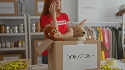 Woman organizing donations in charity room with stuffed animals in box, wearing volunteer shirt, talking on phone, surrounded by clothing and supplies in indoor setting.